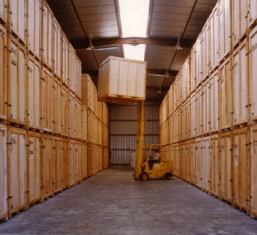 Un chariot élévateur jaune manœuvre un grand container de garde-meuble en bois dans l'allée centrale d'un vaste entrepôt de stockage industriel.