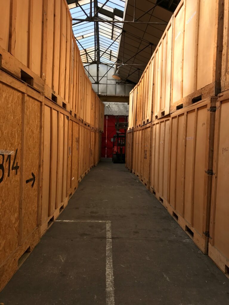 Allée centrale d'un grand entrepôt de garde-meuble sous verrière avec des box de stockage en bois alignés et empilés sur deux niveaux.