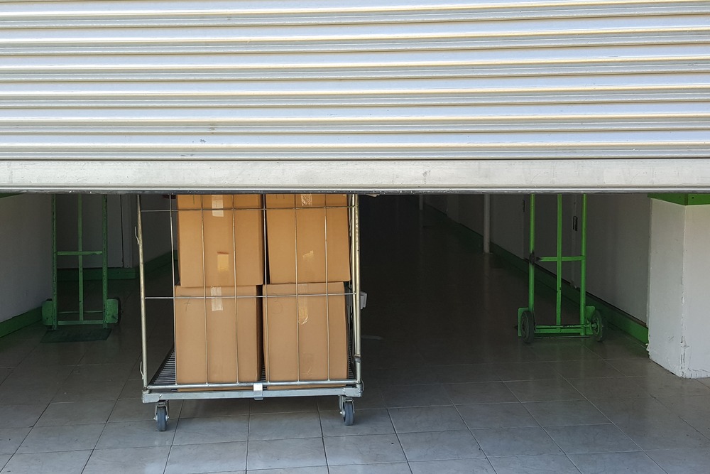 Un chariot métallique grillagé chargé de cartons bruns positionné sous le rideau métallique à moitié ouvert d'un entrepôt de stockage.