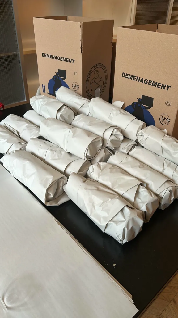 Rouleaux de vaisselle et objets fragiles emballés individuellement dans du papier journal blanc neutre disposés devant des cartons de déménagement ouverts.