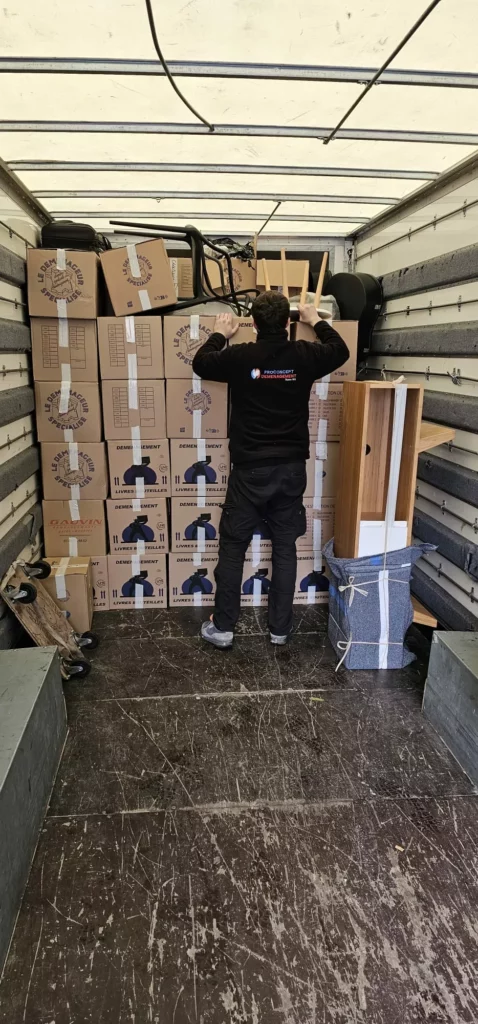 Un déménageur de dos calant une chaise au sommet d'une pile de cartons optimisée à l'intérieur d'un camion capitonné.