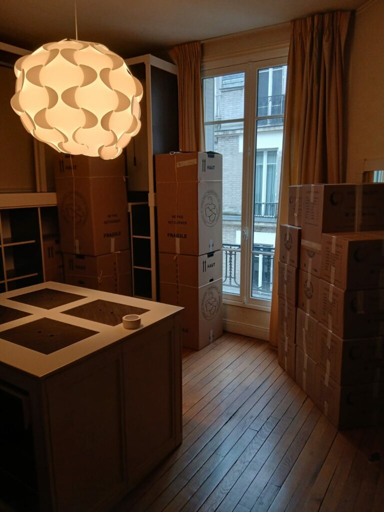 Piles de cartons de déménagement empilées dans une pièce à côté d'un îlot central sous un grand luminaire design suspendu.