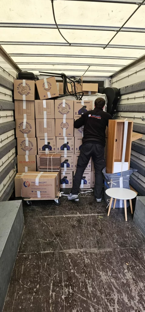 Un déménageur de dos optimisant le rangement de piles de cartons et de mobilier sanglé à l'intérieur d'un camion capitonné.