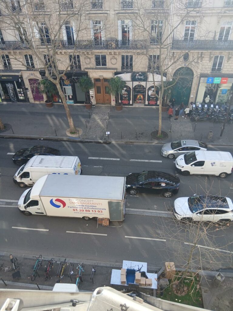 Vue aérienne plongeante depuis une fenêtre sur un camion Proconcept garé le long d'un boulevard parisien bordé d'arbres lors d'un déménagement.