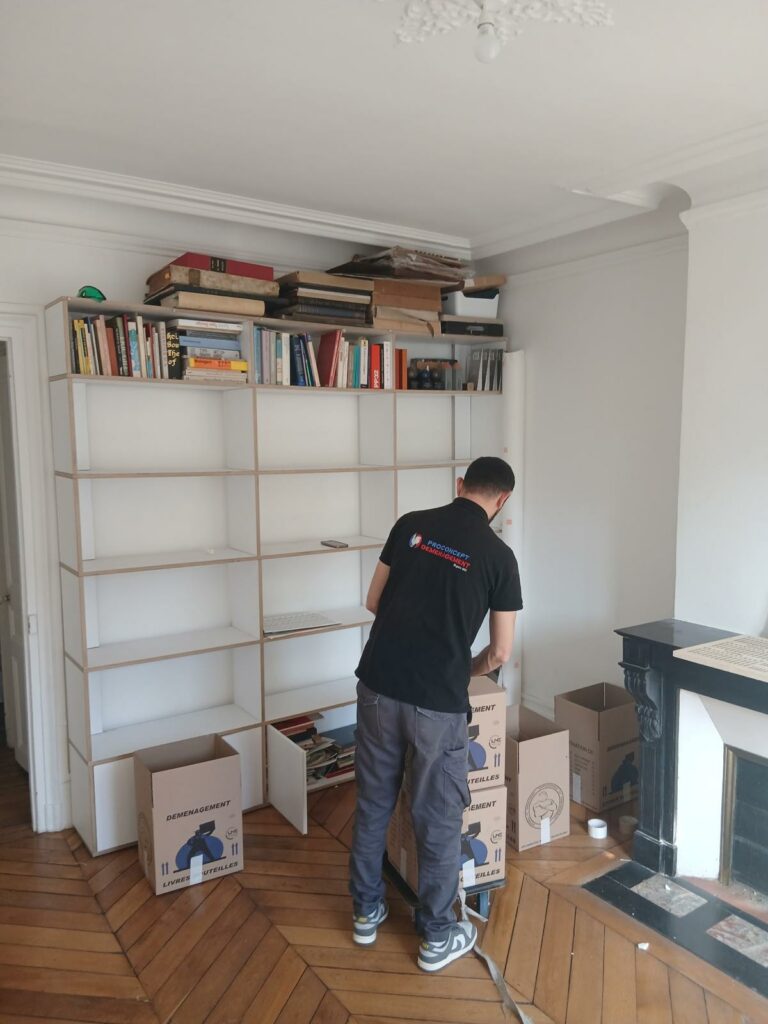 Un déménageur Proconcept met en carton les livres d'une grande bibliothèque blanche dans un salon au parquet ancien.